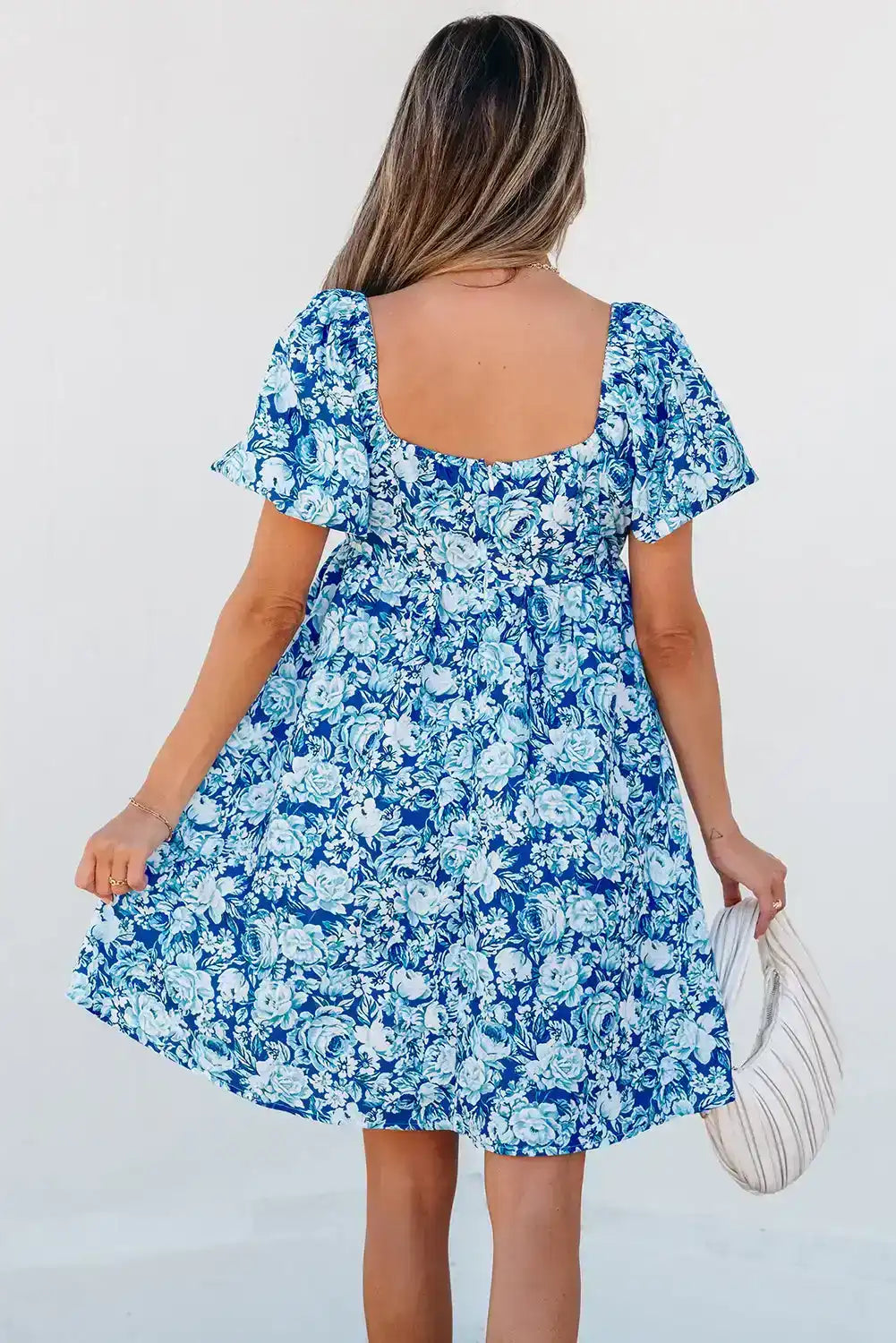 Woman in short sleeve blue floral dress holding a white handbag, seen from the back