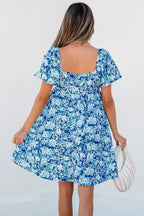 Woman in short sleeve blue floral dress holding a white handbag, seen from the back