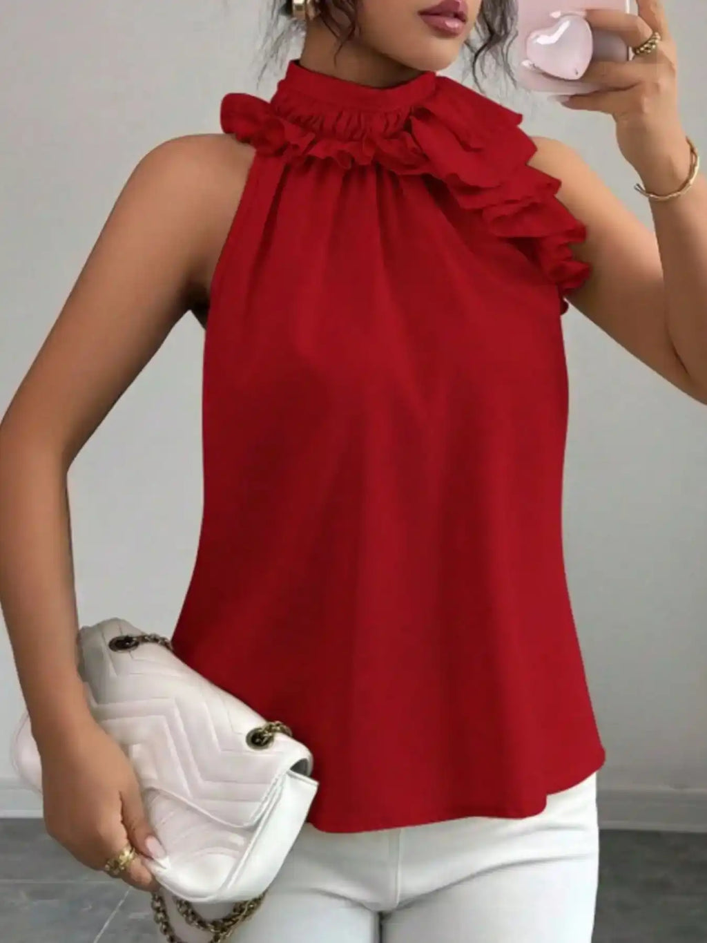 Woman wearing red ruffle neck sleeveless blouse, white pants, and holding a quilted clutch