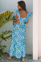 Woman wearing a blue floral maxi dress with ruffle sleeves, standing outdoors by a plant