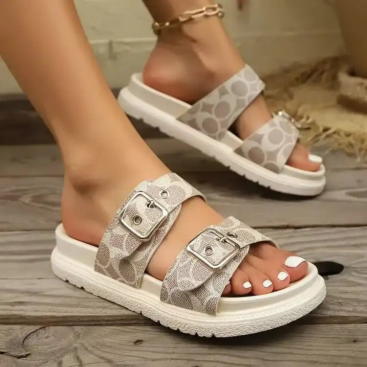 Women’s beige double buckle slide sandals with patterned straps and white soles on wooden floor
