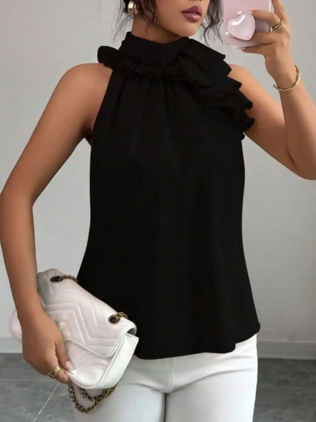 Woman wearing sleeveless black ruffle blouse with white pants and holding quilted clutch