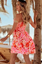 Woman in a sleeveless pink floral summer dress and jewelry on a beach with palm trees.