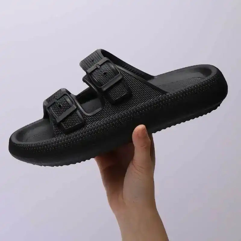 Black slide sandal with double buckle straps for women, held by a hand against a plain background.