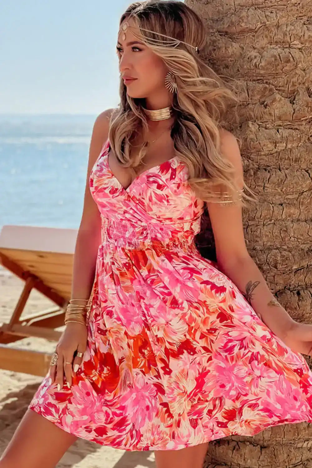 Woman in a pink floral summer dress with gold jewelry on a beach by the ocean.