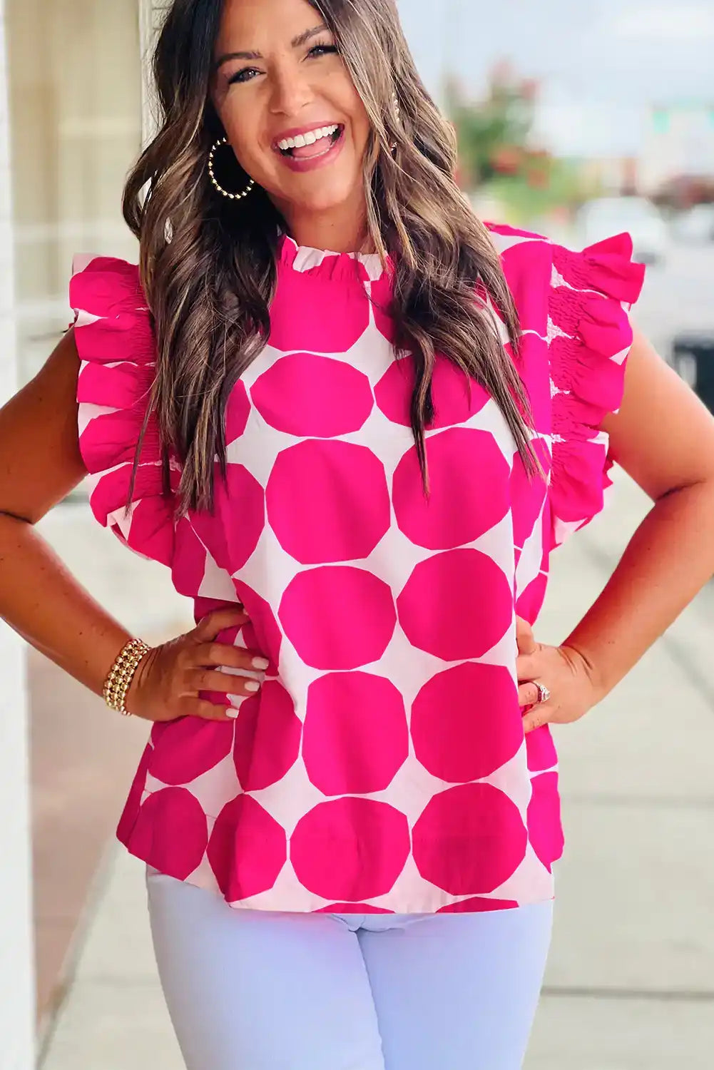 Woman wearing a pink polka dot ruffle sleeve blouse and white pants, smiling outdoors.