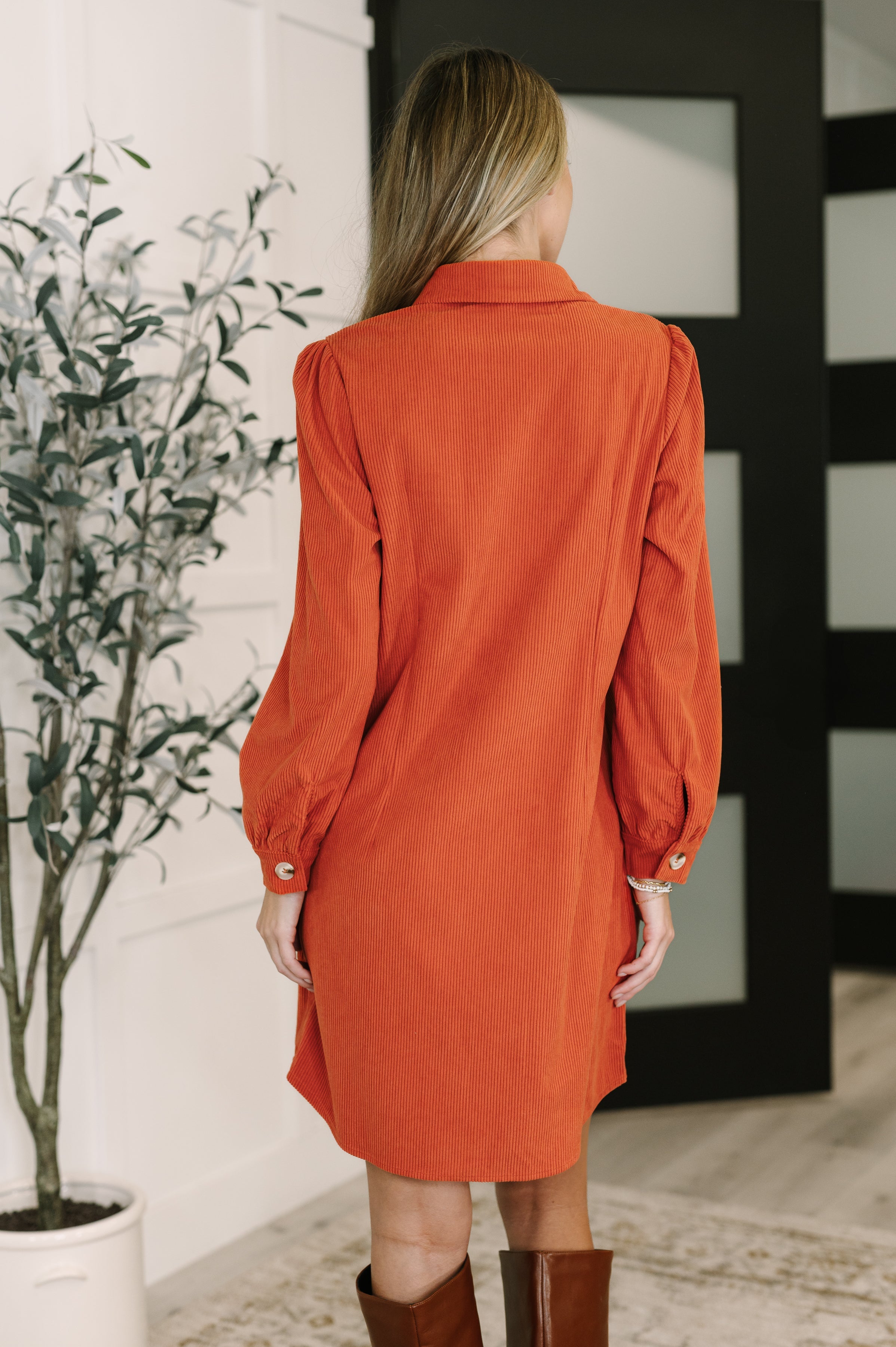 Person wearing an orange dress standing indoors next to a plant.