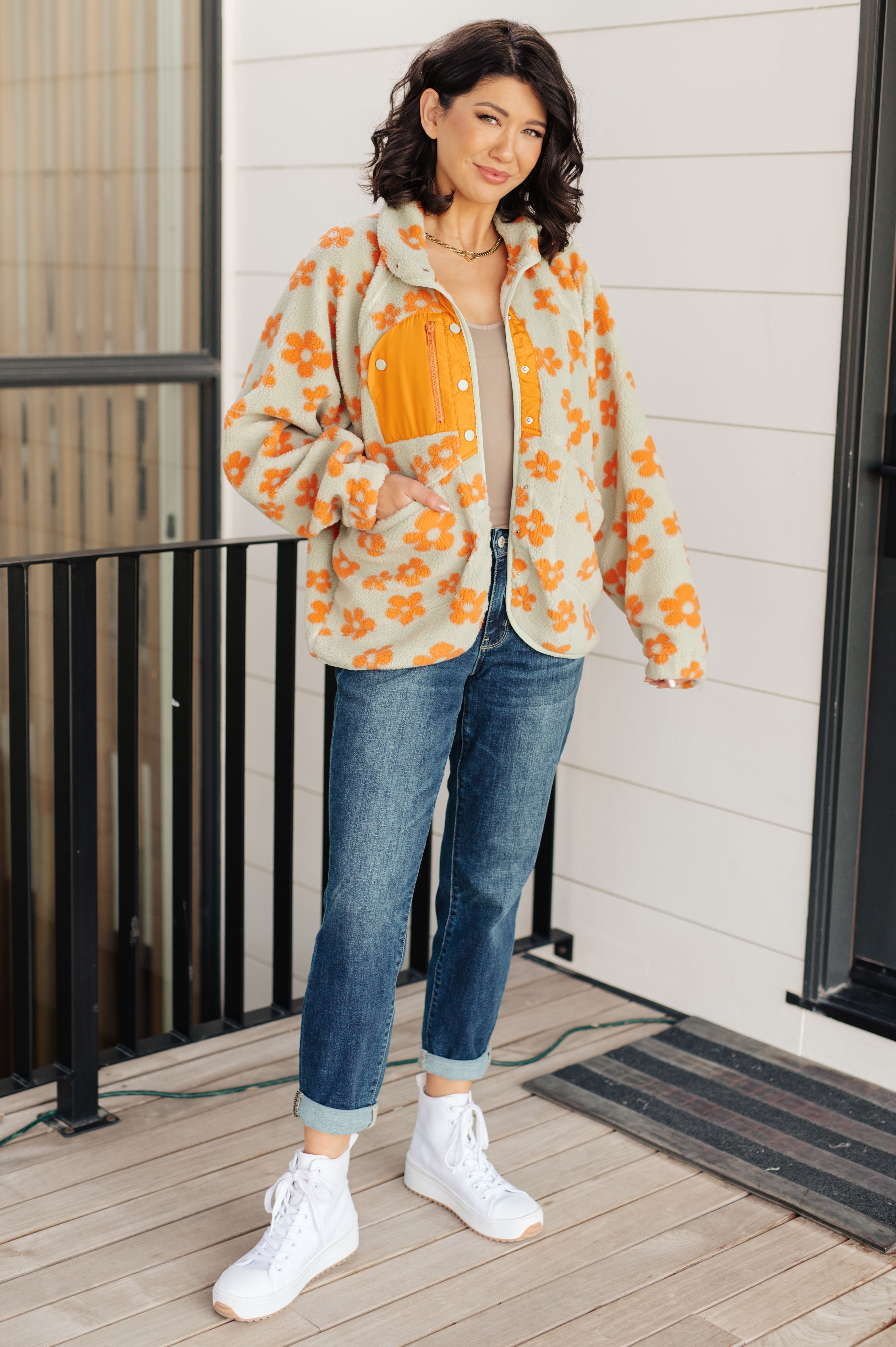 Woman wearing a floral jacket with orange flowers, white shirt, blue jeans, and white sneakers on a wooden deck.