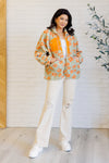 Woman wearing a floral jacket with orange flowers, white pants, and white shoes in a room with a white brick wall and wooden floor.