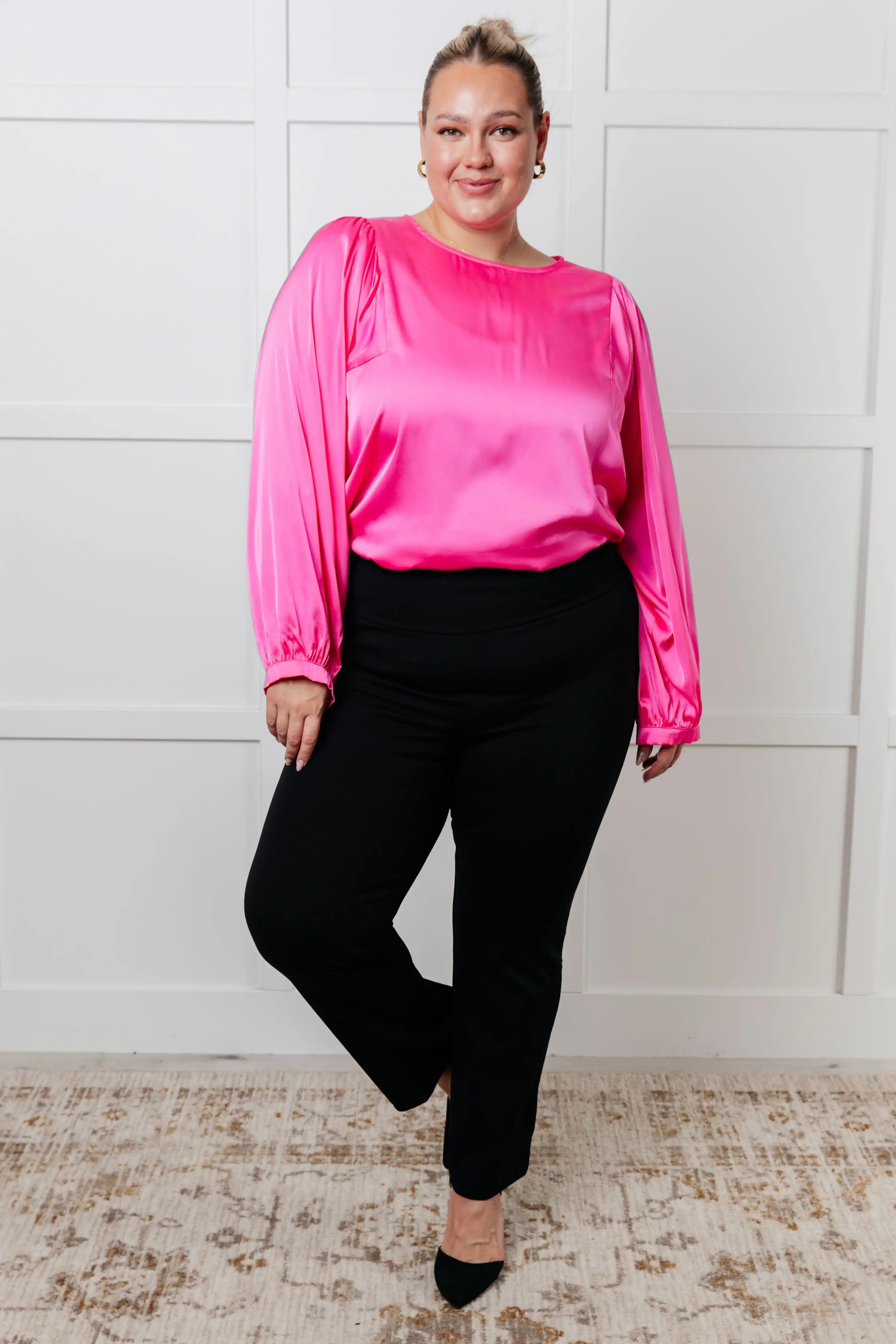 Woman wearing a bright pink blouse and black pants against a white paneled wall.