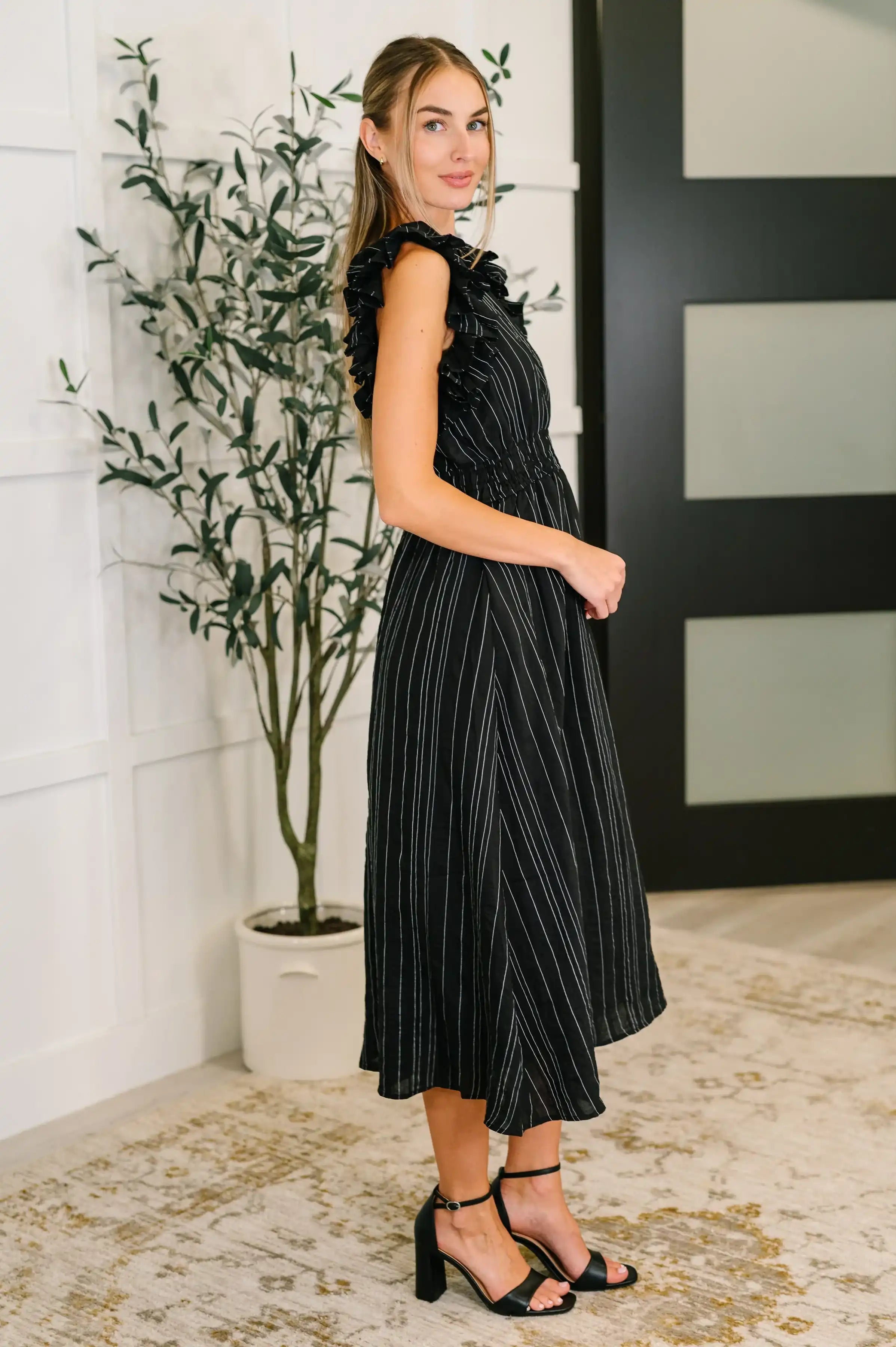 Woman wearing a black pleated dress standing indoors with a plant in the background