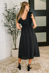 Woman wearing a black ruffled dress standing in a room with a plant and tiled wall.