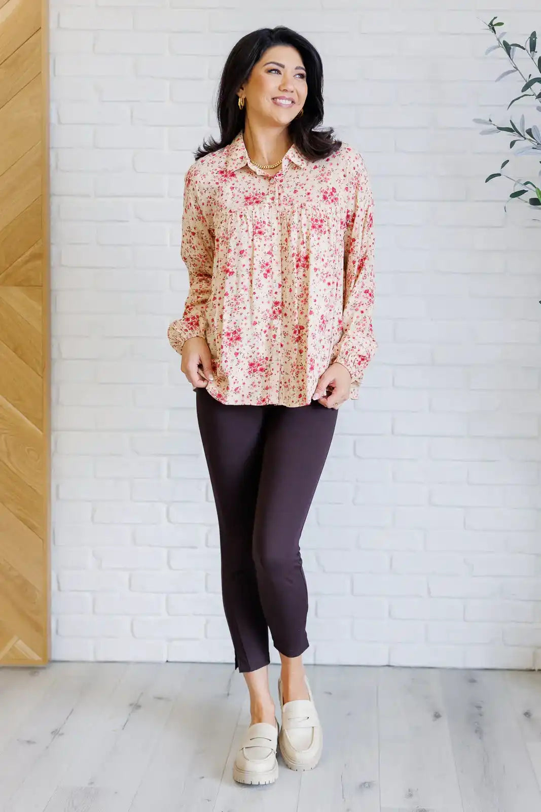 Woman wearing a floral blouse and dark pants against a white brick wall.
