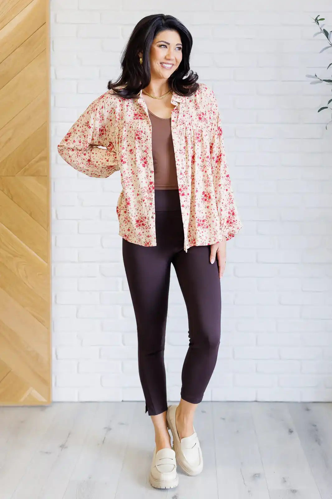 Woman wearing a floral blouse, brown top, and dark pants against a white brick wall.