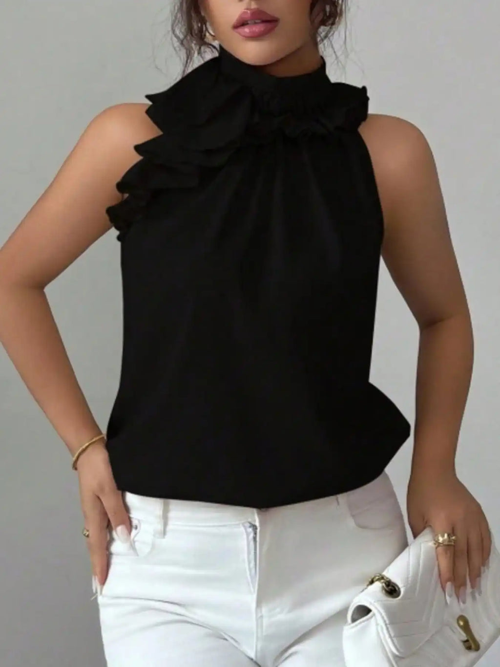 Woman wearing sleeveless black ruffle blouse with high neck and white jeans, holding white clutch
