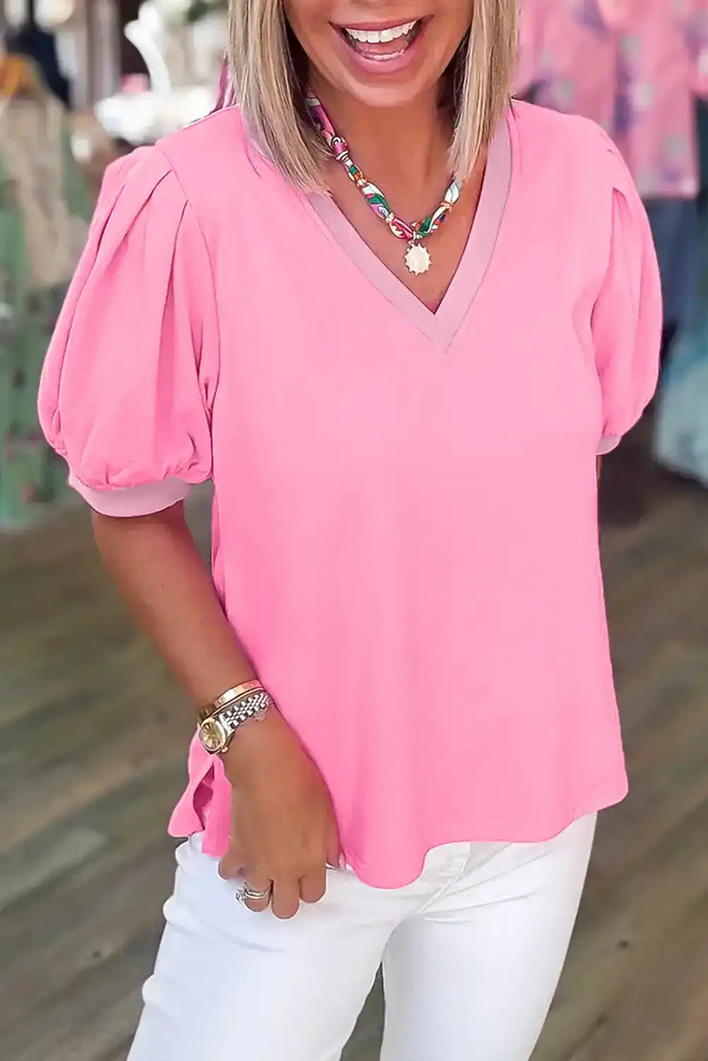 Woman wearing a pink puff sleeve V-neck blouse with white pants and colorful necklace